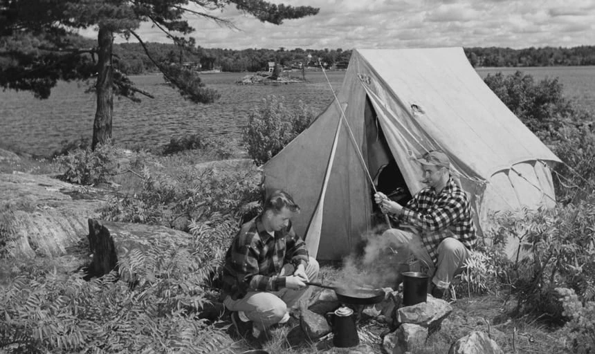 Two men outside camping, one with fishing pole one cooking, next to lake and tent - black and white vintage