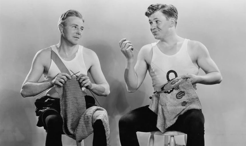 Two men sitting down and knitting while looking at each other and wearing white singlets - black and white vintage
