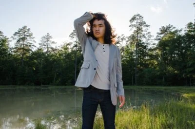 Male model Wearing TravelSmith On The Fly Lightweight Year Round Travel Blazer by Territory Ahead Outside In Park While Holding Hair and Looking Into The Distance