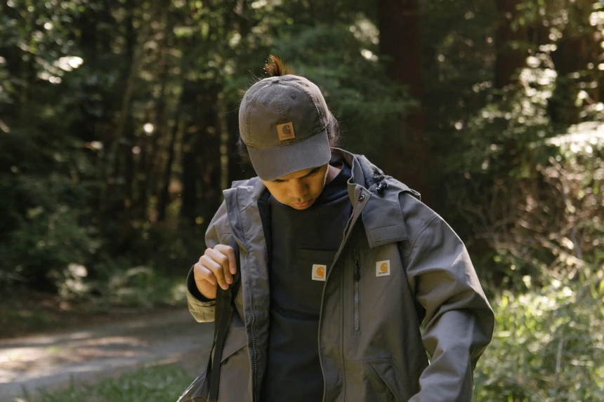 Man in woods holding backpack looking down wearing Carhartt Odessa Cap Rugged Flex Rigby Dungaree and Force Delmont Tee