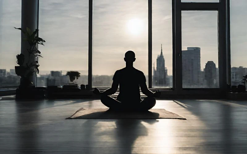 Silhouette image of a man sat meditating in front of a large window view of a cityscape